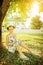 Summer education background: cute little girl sitting on grass and writing in notebook under the tree in warm sun rays. House and