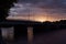 Summer evening with sunset sky, golden sunbeams, blue clouds on promenade, black silhouette bridge with lanterns lights over river