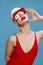 Summer fashion shooting. A sophisticated red-lipped lady in a swimming cap and a red swimsuit touches her glasses and