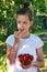 in summer garden young sweet girl in white T-shirt holds a glass vase with ripe cherries and brings one berry to her mouth.