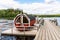 Summer lake nature landscape view of a traditional Scandinavian water floating red wooden sauna spa next to a jetty.