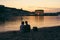 Summer romantic atmosphere: couple in love sits on the river bank at sunset