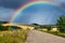 Summer rural landscape - road with cyclists in the rays of the setting sun on background distant rainbow