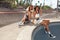 Summer. Skater Girls Sitting On Concrete Skate Ramp At Skatepark. Female Hipsters In Casual Outfit With Skateboard.