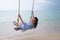 Summer solar portrait of fashion of a way of life of the young stylish woman,sitting on a swing on the beach,carrying lovely fashi