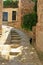 Summer sunny afternoon, a cozy courtyard with yellow stone walls of houses, a black cat walks along the stone steps along a narrow