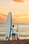 Summer surfing. Father and daughter standing at the beach with a sup board. Ocean and the sunset in the background. Back