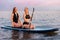 Summer surfing. Two beautiful women swimming on a sup board. In the background, the ocean and pink sunset. Summer active leisure