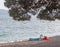 Summer vacation alone, relax. The girl lies on a deserted pebble beach at the edge of a calm sea under the branches of a pine wrap