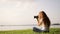 Sun-kissed freckled redhead girl with photo-camera is sitting on a green grass and takes a photo in forest near the lake