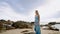 Sun-kissed woman strolls on sandy beach in blue dress. Female enjoys serene sea, rocks backdrop. Leisure, vacation