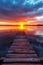 The sun sets in a blaze of color as a weathered pier leads into tranquil waters, reflecting the dramatic sky.