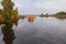 The sun shines slightly through the clouds. The quiet water of a river or lake glistens with reflections. Yellow reeds and trees.