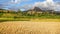 Sun shines on wheat fields in foreground, green and yellow rice terrace fields on small hills, clay houses and rocky mountain