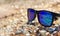 Sunglasses with blue lenses lie on a sandy shell beach on the sea front in sunny weather.