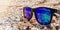 Sunglasses with blue lenses lie on a sandy shell beach on the sea front in sunny weather. Recreation and tourism concept