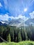 Sunlit mountain range, with lush green trees and snow-capped peaks in the background