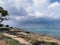 Sunny coast of the Mediterranean Sea with trees and stones, dramatic sky and rain above the sea, boats in the sea and the largest