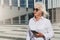 Sunny day.Middle-aged woman,retired woman dressed in white shirt and sunglasses,stands on city street,holds smartphone