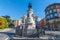 Sunny warm day. Statue of Maria Christina of Borbon in Madrid, Spain. People walking along sidewalk.