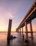 Sunrise casting golden light on a long bridge crossing a body of water. Rocks and old pilings in the foreground