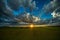 Sunset in field with fence, green pasture and cattle under blue sky with clouds and penetrating sunbeams