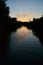 Sunset on a narrow river: silhouetted, shadowed trees, clouds, golden sky reflected in water