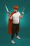 Super hero handyman holding saw and hammer. Man wearing red helmet, mask and cape holds building equipment standing on