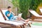 Suprised bearded young adult freelancer man in blue t-shirt and shorts lying on deckchair on poolside, holding hands on cheeks,