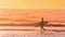 Surfer boy with his surfboard walking along the beach at sunset