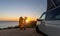 Surfer boy sitting near his mini van and looking on the ocean at summer sunset with a surfboard on her side