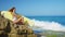 Surfer girl sits on a beautiful rocky beach with board. Powerful waves hit the rocky shore. Girl looking into distance.