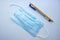 A surgical or medical mask with rubber ear straps lies next to a pen and pencil on a white background. A typical 3-layer doctor`s