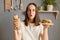Surprised amazed woman with dark hair eating delicious burger and hot dog, breaking diet and likes fast food, wearing white T-