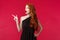 Surprised and astounded young redhead woman in elegant party black dress, drop jaw gaspins as staring and pointing