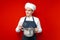 Surprised chef holds dishes and prepares a dish on a red isolated background, kitchen worker in uniform with kitchen items