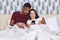 Surprised dark skinned man embraces his wife, pose together in comfortable bed, view photos or read news in internet on cell phone