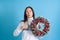surprised, delighted Asian young woman holds Christmas wreath and thumbs up isolated on blue background