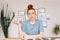 Surprised disappointed redhead young woman student is sitting at a table with books and a workbook.