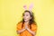 Surprised emotional curly little girl wearing bunny ears isolated on yellow background. Easter holiday concept