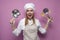 Surprised girl cook in apron holds dishes and kitchen items on a pink background, portrait of a housewife woman