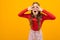 Surprised girl with curly hair in a red blouse and striped trousers peers through the palms on an orange background with