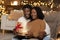 Surprised glad millennial african american lady and man opens box with gift, light in cozy living room
