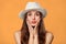 Surprised happy beautiful woman looking sideways in excitement. Excited girl in hat, isolated on orange background