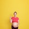 Surprised pregnant woman pointing finger up isolated over yellow background, lady with widely opened mouth touching her bare tummy