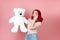 surprised, shocked young woman with open mouth and red hair holds large white teddy bear isolated on a pink background