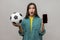 Surprised woman holding and showing blank display smartphone and football ball, betting on soccer.