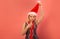 Surprised woman holds Santa Claus hat, wears silver dress with Christmas tinsel and looks at camera on pink background.