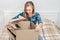 Surprised young woman sitting on bed unpacking cardboard box at home. Delivering parcel