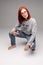 Swag and confident red haired girl in jeans and sweatshirt posing at gray studio.